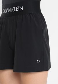 Black Calvin Klein shorts with elastic waistband featuring brand name in white, small CK logo near hem, worn on lower torso and upper thighs.