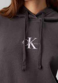 Felpa grigia con cappuccio e cordini, con logo ricamato bianco "CK" e testo "Calvin Klein Jeans" sul davanti. Texture morbida.