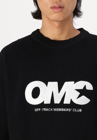 Jersey negro acanalado con cuello redondo y un llamativo texto bordado en blanco "OMC" con "OFF-TRACK MEMBERS' CLUB" debajo en texto más pequeño.