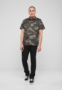 Korte mouw camouflage shirt in groen- en grijstinten, voorzien van twee borstzakken, zwarte knopen, gecombineerd met zwarte jeans en sneakers.