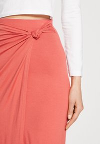 Rok fabric coral dengan detail simpul di pinggang dan desain berpola. Tekstur lembut, panjang sedang, dipadukan dengan atasan lengan panjang putih.