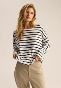 Czarnobiały pasek długim rękawem z dekoltem w łódkę; ma małe złote logo na piersi i luźny krój, połączony z beżowymi spodniami.