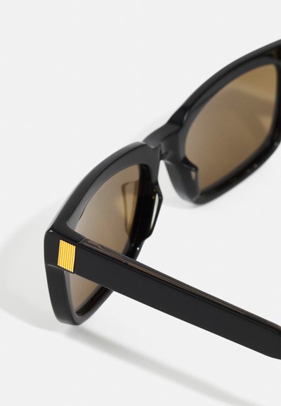Schwarze Sonnenbrille mit braunen Gläsern, ausgestattet mit einem markanten Rahmen, einem leichten geometrischen Akzent und einem strukturierten gelben Streifen am Bügel.