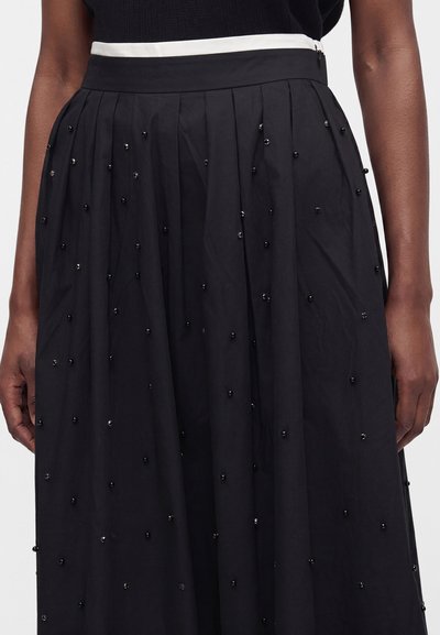 Jupe plissée noire avec de petites perles noires dispersées sur le tissu et une ceinture blanche portée par une personne à la peau foncée.