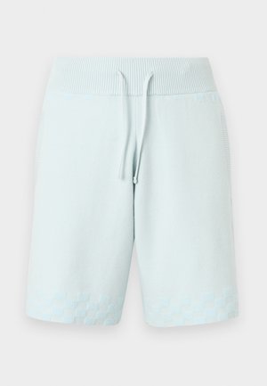 Mann trägt ein weißes offenes Hemd über einem weißen Tanktop, hellblaue Shorts, cremfarbene Socken und zweifarbige Loafers, steht mit einer Hand in der Hosentasche.