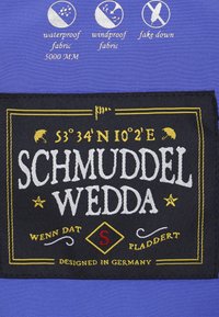 Blå vattentät och vindtät tyg, med en svart label med vit och gul text, inklusive geografiska koordinater och design detaljer.