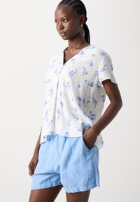 Blusa floreale bianca con motivi blu, maniche corte e scollo a V. Abbinata a pantaloni corti larghi di un azzurro chiaro con tasche.