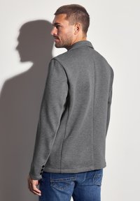 Harmaa bleiseri, jossa on teksturoitu neulekangas, varustettuna kauluksella, pitkillä hihoilla ja kolmella mustalla napilla hihassa, päällä siniset farkut.