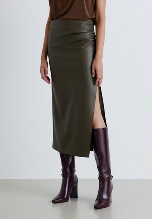 Donna che indossa una gonna midi in pelle verde scuro con uno spacco laterale e stivali al ginocchio con tacco viola scuro, in piedi davanti a uno sfondo semplice.