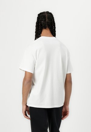 Person mit langen, verdrehten Zöpfen, der ein einfaches weißes Kurzarm-T-Shirt und schwarze Hose trägt und mit dem Rücken zu sehen ist vor weißem Hintergrund.