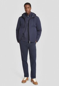 Giacca imbottita blu navy con cappuccio, sovrapposta a un top e pantaloni coordinati. Scarpe marroni con lacci bianchi. Texture liscia e design aderente.