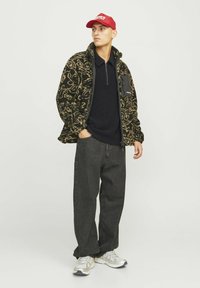 Veste en polaire à motifs noir et beige avec une poche zippée, associée à un polo noir et un jean gris foncé. Le mannequin porte une casquette rouge et des baskets.