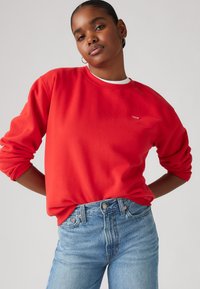 Red crew-neck sweatshirt with long sleeves and cuffed hems, featuring a small logo on the chest. Paired with light blue high-waisted jeans.