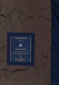 Marine stof med en glat tekstur, som har et rektangulært label med teksten "Vandafvisende" fra Hackett, med gyldne syninger som accent.