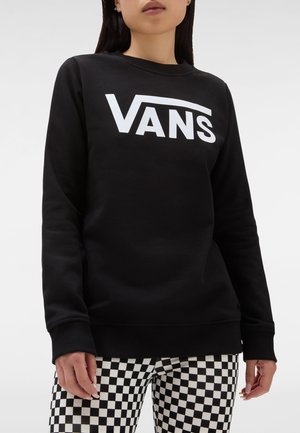 Kobieta ubrana w czarną bluzę z białym logo Vans oraz czarno-białe spodnie w kratkę na jednolitym jasnym tle.