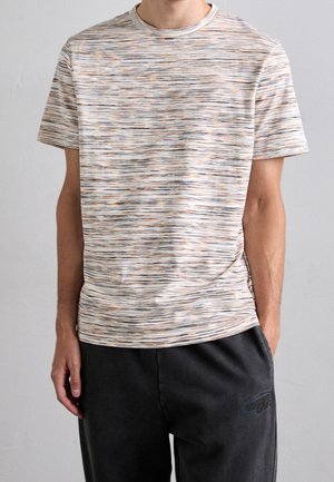 Mann trägt ein kurzärmeliges, mehrfarbig gestreiftes T-Shirt und schwarze Hose mit einer Hand in der Tasche vor hellem, einfarbigem Hintergrund.