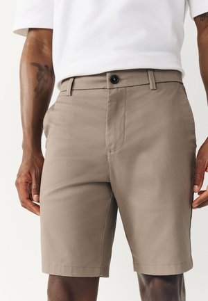 Homme portant un short beige sur mesure et une chemise blanche, montrant un gros plan du short avec un bouton et des poches, ainsi qu'un tatouage sur l'avant-bras gauche.