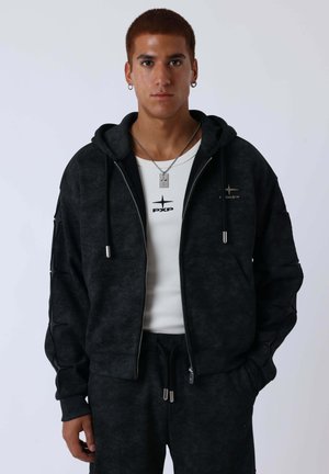 Jonge man met kort haar die een zwarte hoodie met ritssluiting draagt, bijpassende zwarte joggingbroek, wit T-shirt met logo en een kruis ketting, staand en naar voren kijkend.