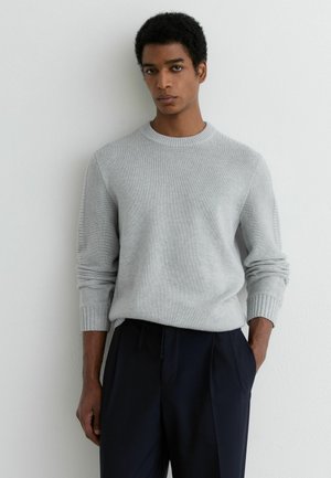 Mężczyzna w jasnoszarym swetrze z dzianiny z prążkowanymi mankietami i granatowych spodniach, stojący na tle gładkiej białej ściany, zwrócony przodem.