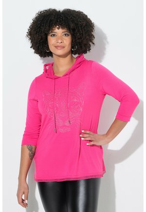 Kvinde med krøllet hår iført en knaldpink hættetrøje med en tigeransigt af rhinsten, sorte leggings og store øreringe, stående med hånden på hoften.