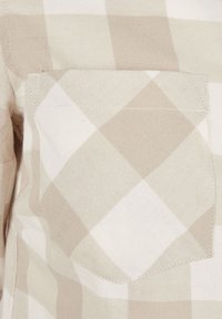 Tissu à carreaux beige et blanc avec une poche poitrine. Présente une texture tissée en diagonale et des coutures contrastantes sur le bord de la poche.