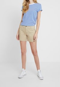 Stribet blå og hvid t-shirt med opkrogede ærmer, kombineret med beige shorts. Modellen bærer hvide høje sneakers. Ensfarvet hvid baggrund.