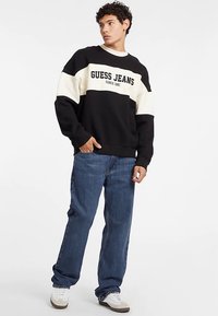 Zwart-witte sweatshirt met de tekst "GUESS JEANS", oversized pasvorm, gecombineerd met blauwe recht uitlopende jeans en witte sneakers. Gladde textuur.
