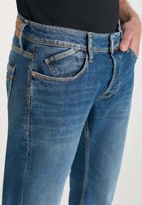 Jeans in denim blu con vestibilità regolare, cinque tasche e lieve scolorimento. Presentano una toppa in pelle sulla vita e cuciture in contrasto.