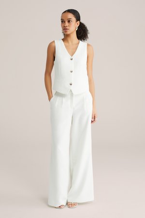 Mujer con cabello rizado, vestida con un chaleco blanco sin mangas y pantalones de pierna ancha, de pie con las manos en los bolsillos sobre un fondo claro.