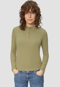 Olijfgroene polo met lange mouwen en een gestructureerd ribbelpatroon, voorzien van een knoopsluiting en een klassieke kraag. Gecombineerd met blauwe jeans.