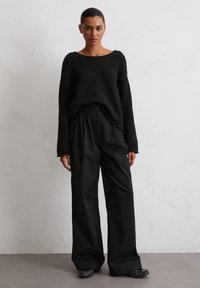 Pull noir, texturé, ample avec des manches longues associé à un pantalon noir taille haute à jambes larges et des chaussures noires à enfiler.