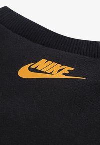 Crna pamučna dukserica s rebrastim izrezom i istaknutim narančastim Nike logom na prednjoj strani, koja ima glatku teksturu.