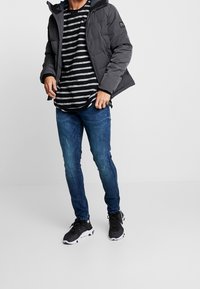 Graue Steppjacke mit Kapuze, getragen über einem schwarz-weiß gestreiften Langarmhemd. Blaue Skinny-Jeans und schwarze Sportschuhe vervollständigen das Outfit.