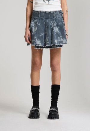 Persona che indossa una gonna di denim plissettata e sfrangiata con grafiche stampate, calzini neri e mocassini neri chunky con testo bianco.