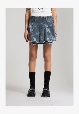 Persona che indossa una gonna di denim plissettata e sfrangiata con grafiche stampate, calzini neri e mocassini neri chunky con testo bianco.