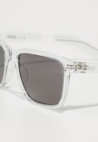 Lunettes de soleil en plastique transparent avec des verres foncés, monture carrée et accents métalliques affichant le logo Puma sur les tempes.