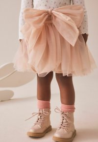 Jupe en tulle rose pâle avec un grand nœud à la taille, associée à des chaussettes rosa claires montantes jusqu'aux genoux et des bottes montantes en dentelle métalliques rose.