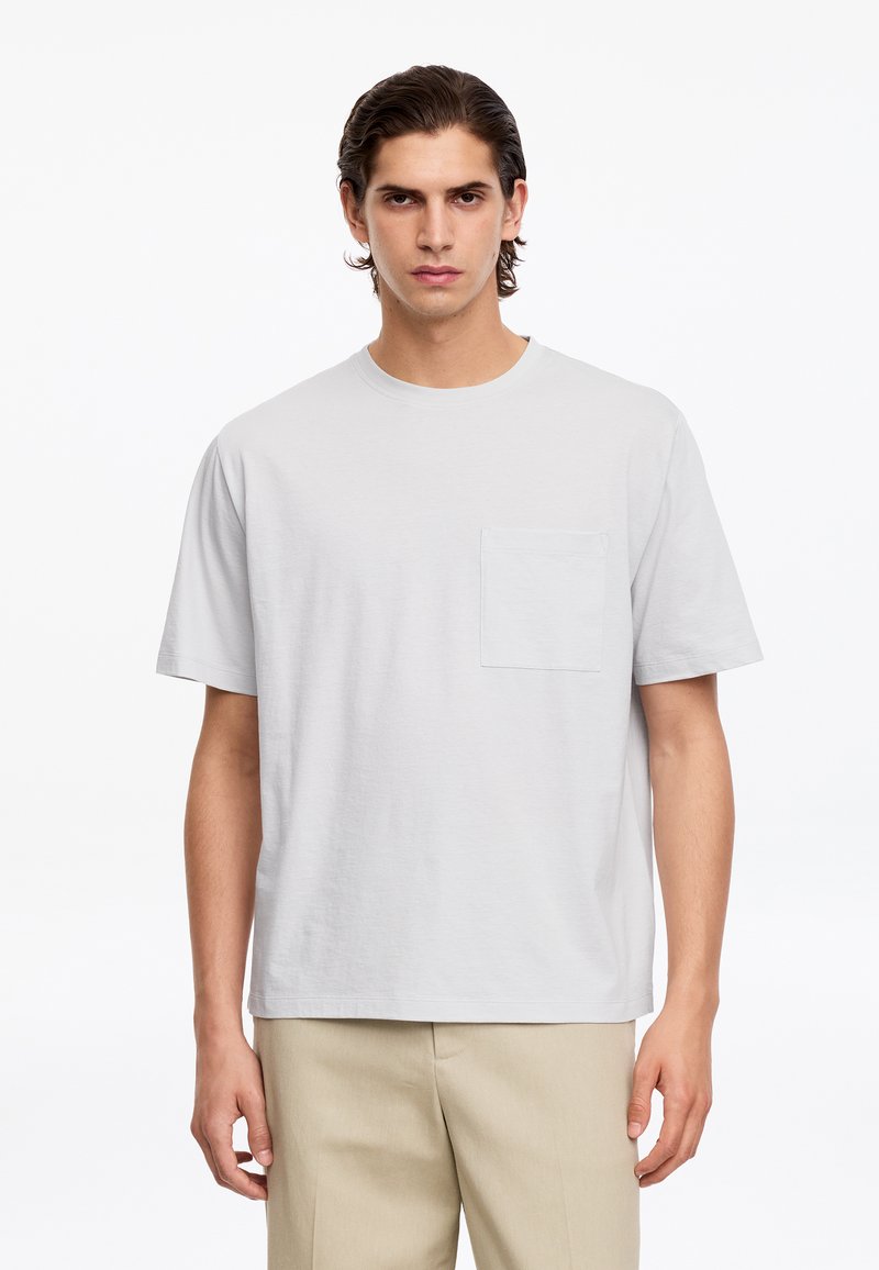 Jeune homme aux cheveux foncés portant un t-shirt blanc uni à manches courtes avec une poche poitrine et un pantalon beige, debout devant un fond blanc.