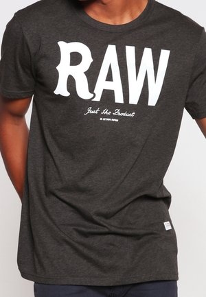Homme portant un t-shirt gris foncé avec un grand texte blanc "RAW" et un texte plus petit "Just the Product G-STAR RAW" sur le devant.