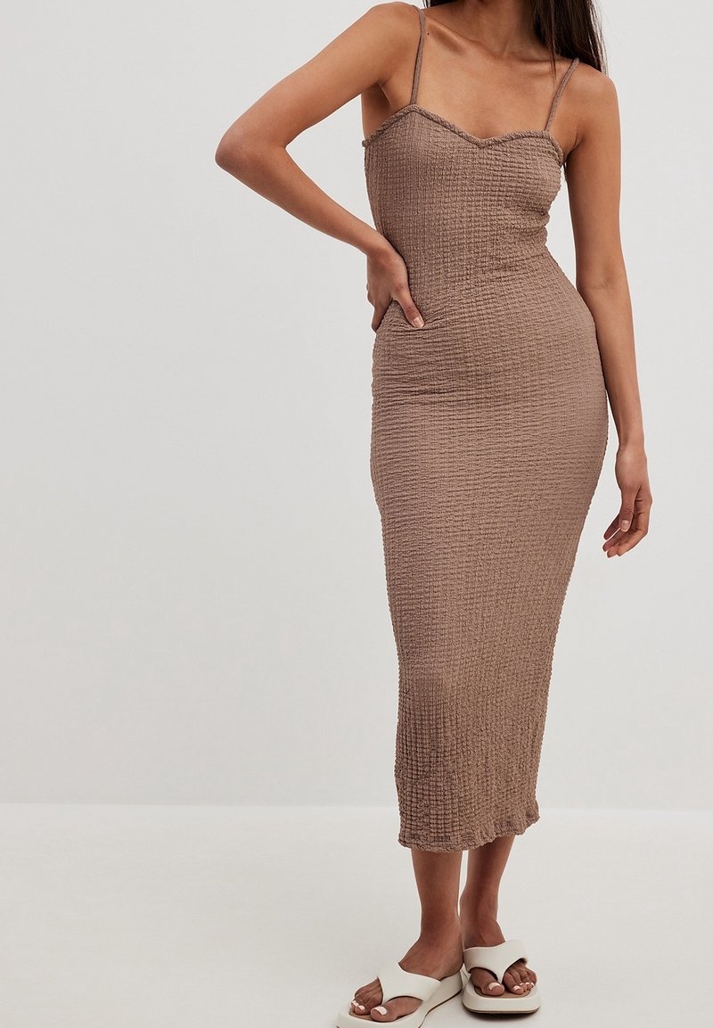 Femme portant une robe midi taupe ajustée et texturée avec de fines bretelles et des sandales blanches à bouts ouverts, debout, une main sur la hanche.