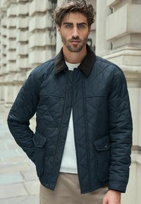Marineblauer gesteppter Blazer mit einem braunen Cordkragen, Reißverschluss vorne und zwei aufgesetzten Taschen; über einem weißen Rundhals-Sweater getragen.