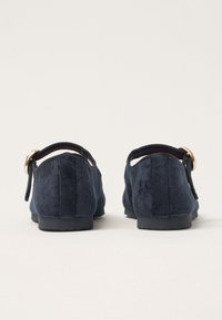 Un par de zapatos planos de terciopelo azul oscuro con suelas negras vistos desde atrás sobre un fondo claro y liso.