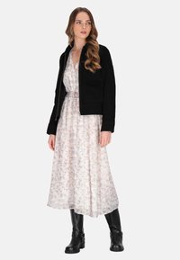 Jachetă bomber din suede negru peste o rochie lungă albă, imprimată cu flori. Modelul poartă cizme negre până la genunchi. Texturi moi și culori în tonuri line.
