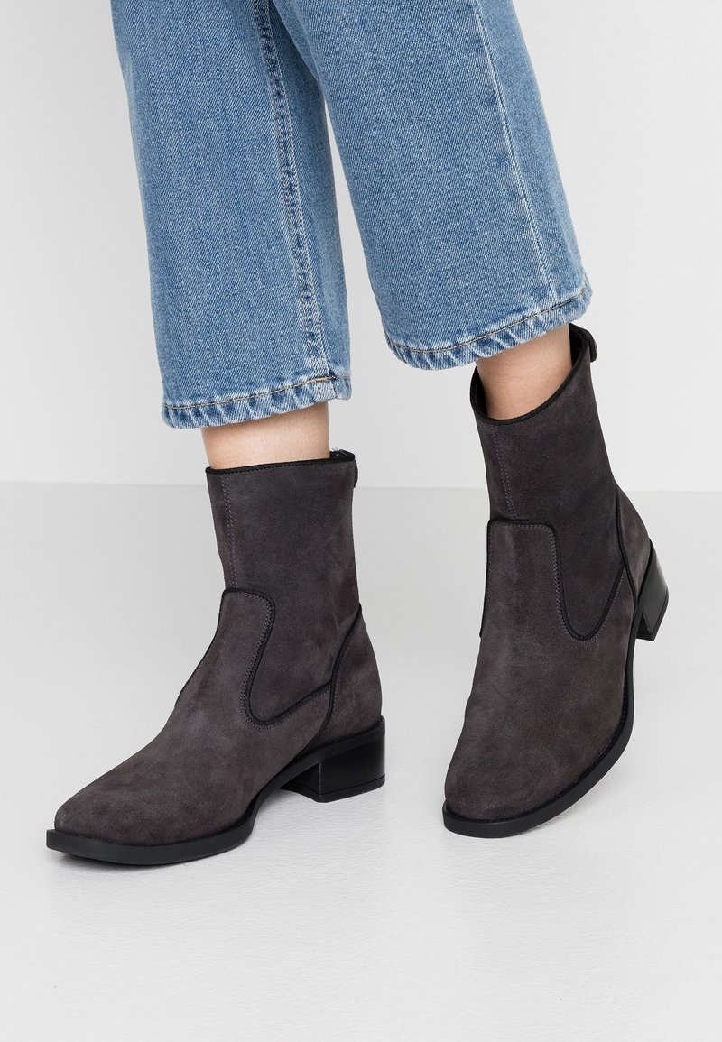 Bottes en daim gris foncé à la cheville avec petits talons portées avec un jean court en denim bleu sur un sol clair.