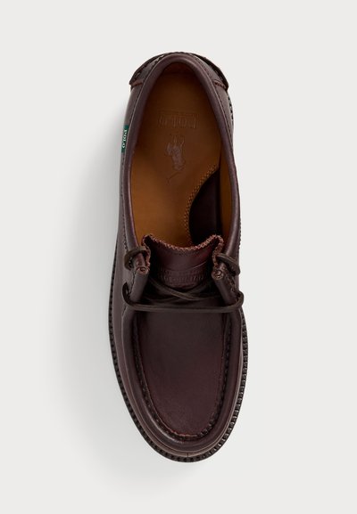 Zapato de cuero marrón con punta redonda, costuras decorativas y diseño con cordones. Presenta una suela marrón texturizada y una marca grabada en el interior.