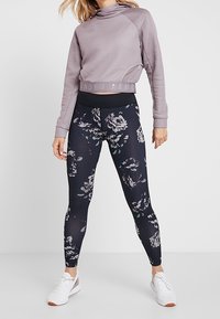 Harmaa urheilullinen crop-top korkean kauluksen ja brändilogo kanssa, yhdistettynä mustiin kukkakuosisin legginsseihin. Materiaali venyy ja malli on kapeneva.