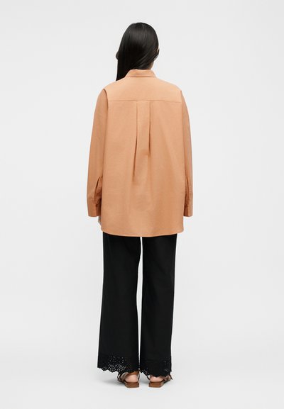 Žena s dlhými tmavými vlasmi, oblečená v voľnej broskyňovej košeli na gombíky, čiernych širokých nohaviciach s krajkovými lemami a hnedých sandáloch, stojaca chrbtom.