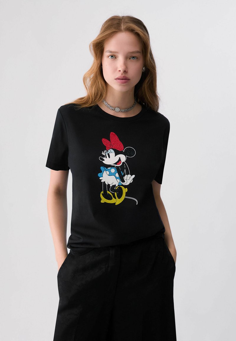 Jeune femme aux cheveux clairs porte un t-shirt noir avec un motif coloré de Minnie Mouse, assorti à un pantalon noir et un collier chaîne argenté.