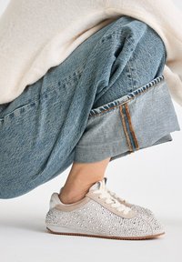 Zapatillas brillantes con parte superior de ante beige, adornadas con piedras de strass transparentes y una suela clara; combinadas con unos vaqueros azules con dobladillo enrollado.