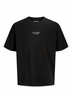 Ragazzo sorridente in piedi, indossa una maglietta nera a maniche corte, pantaloni beige a gamba larga e scarpe da ginnastica bianche su uno sfondo bianco semplice.